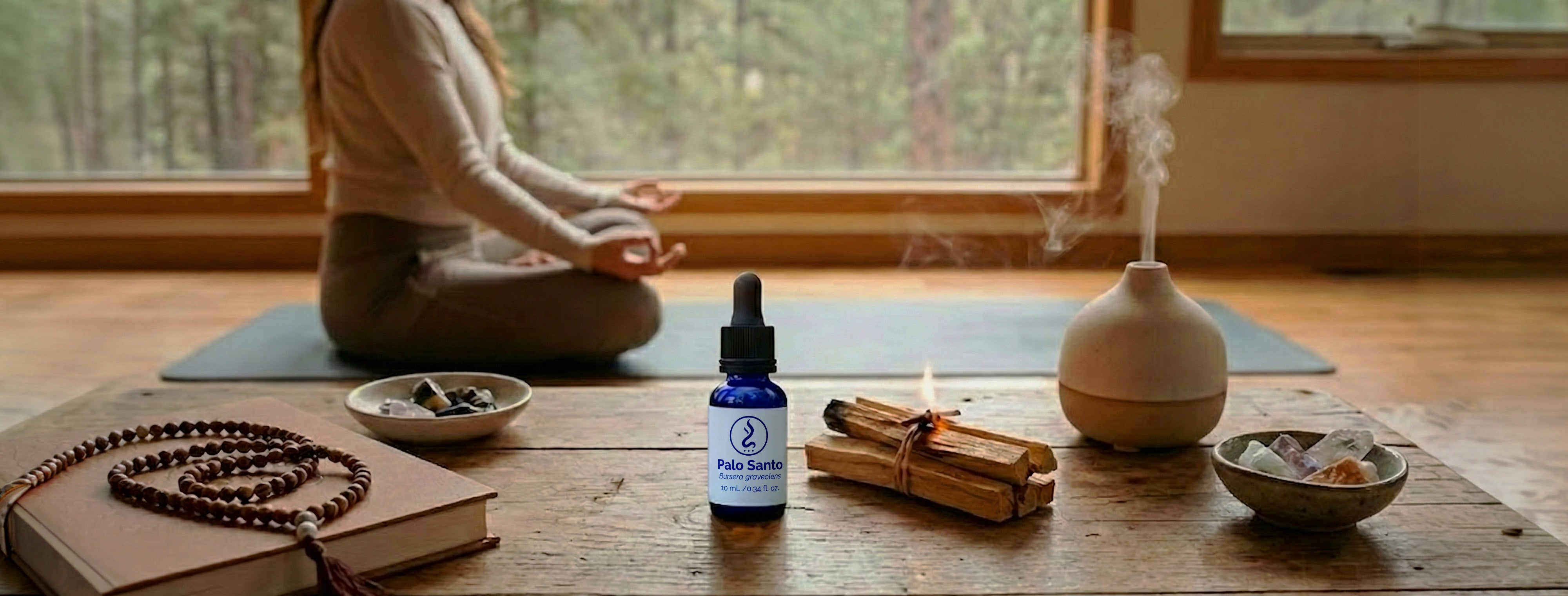 Woman meditating in lotus position by a large window, with Palo Santo oil, burning Palo Santo wood sticks, a diffuser, prayer beads, and crystals on a wooden table in the foreground.