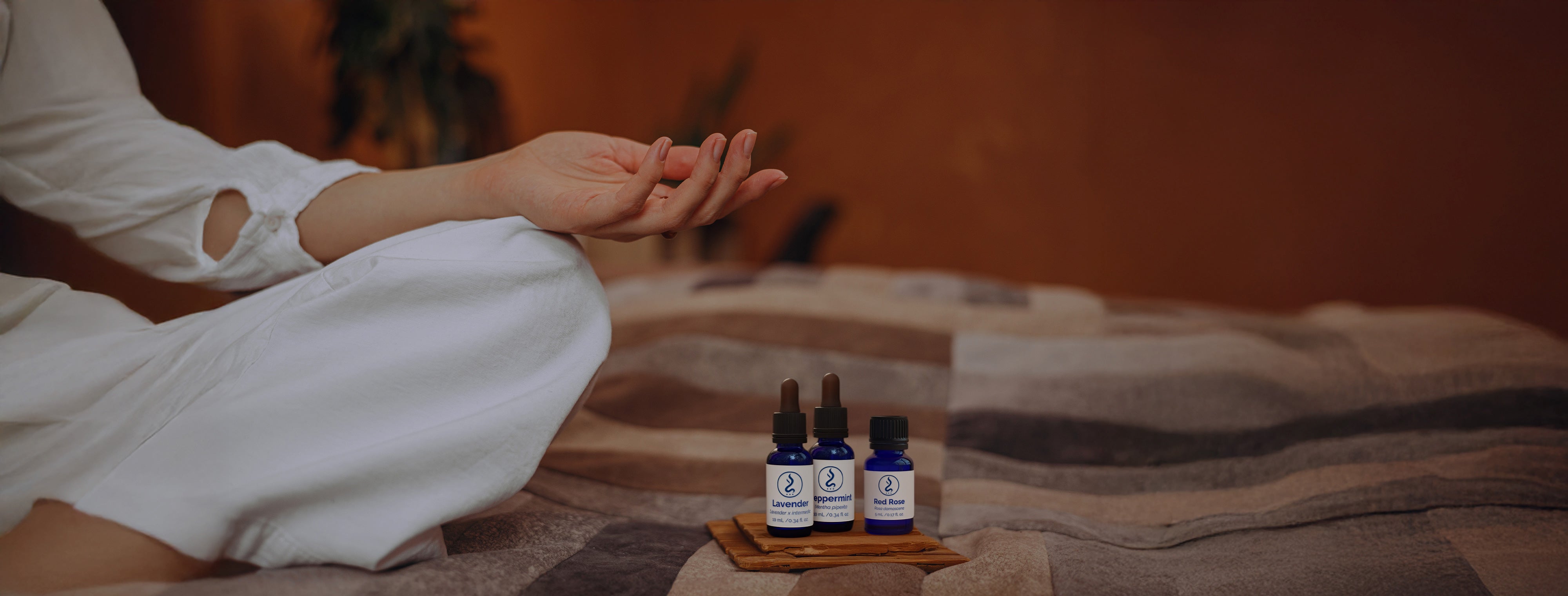 Person meditating in white attire with essential oil bottles placed on a wooden tray nearby.