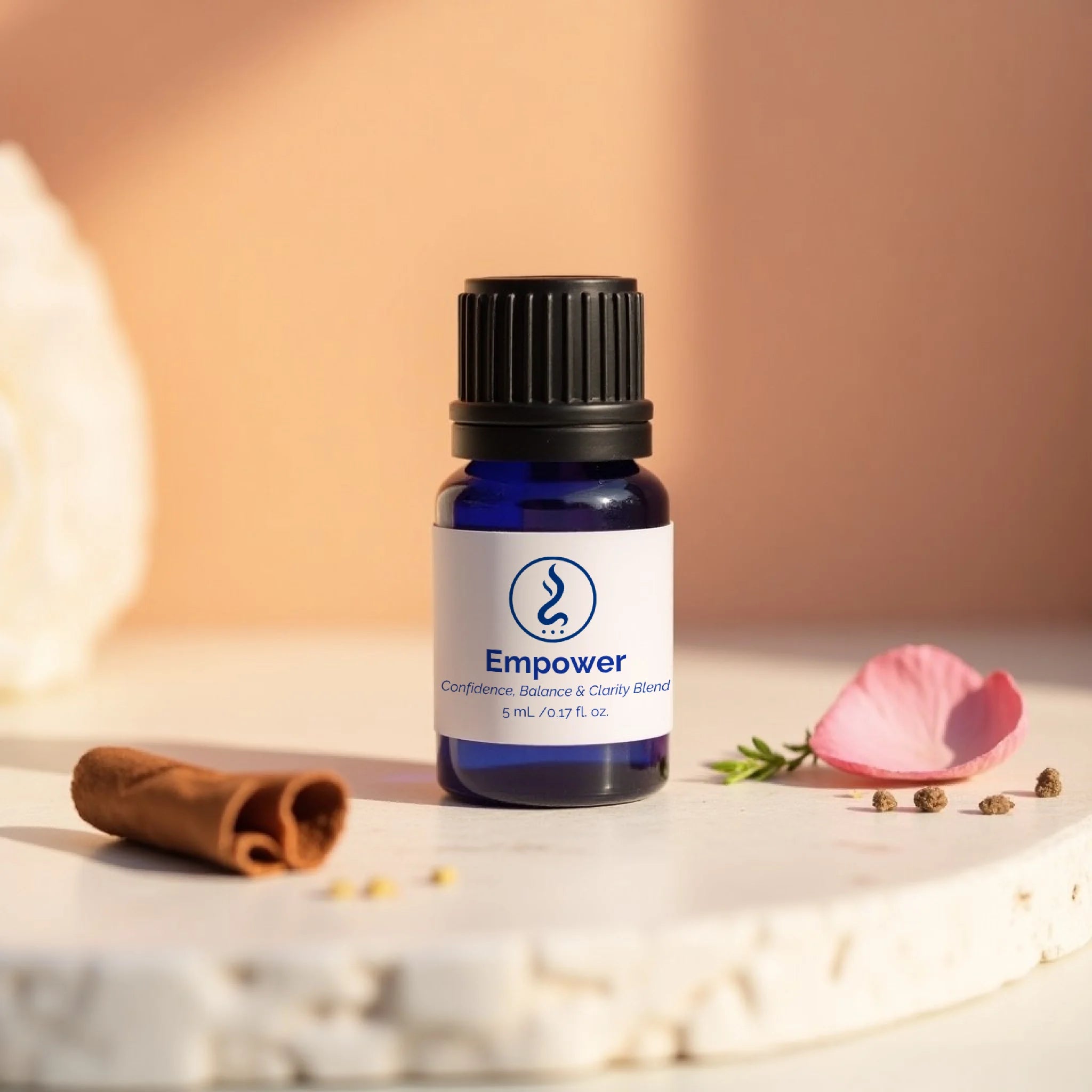 Bottle of 'Empower' sacred oil with a cinnamon stick and pink flower petal on a neutral background